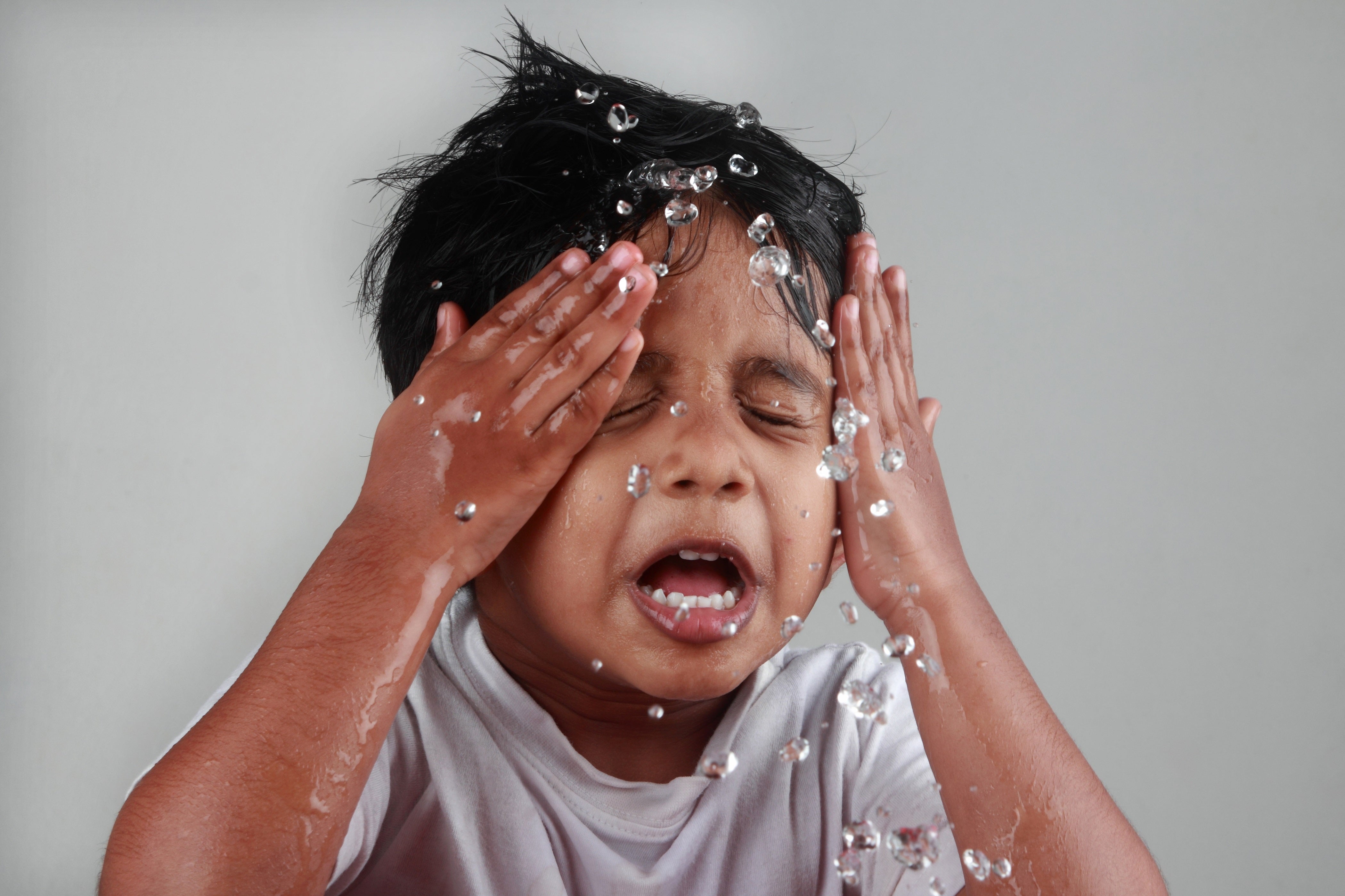 Child splashing water on their face with eyes closed and hands covering their cheeks against a plain background.