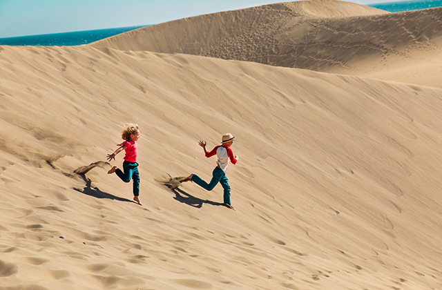 Two children run and play across large sand dunes under a clear sky, exploring an outdoor landscape near the coast. The scene captures adventure, freedom, and active play in nature.