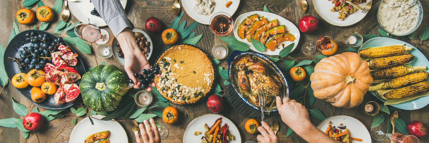 Overhead view of a festive dining table filled with roasted turkey, pumpkins, fruits, and assorted dishes, with people serving and sharing food.