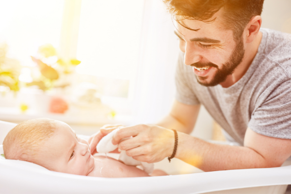 Smiling father gently bathing a baby with a washcloth in a bright and cozy bathroom.