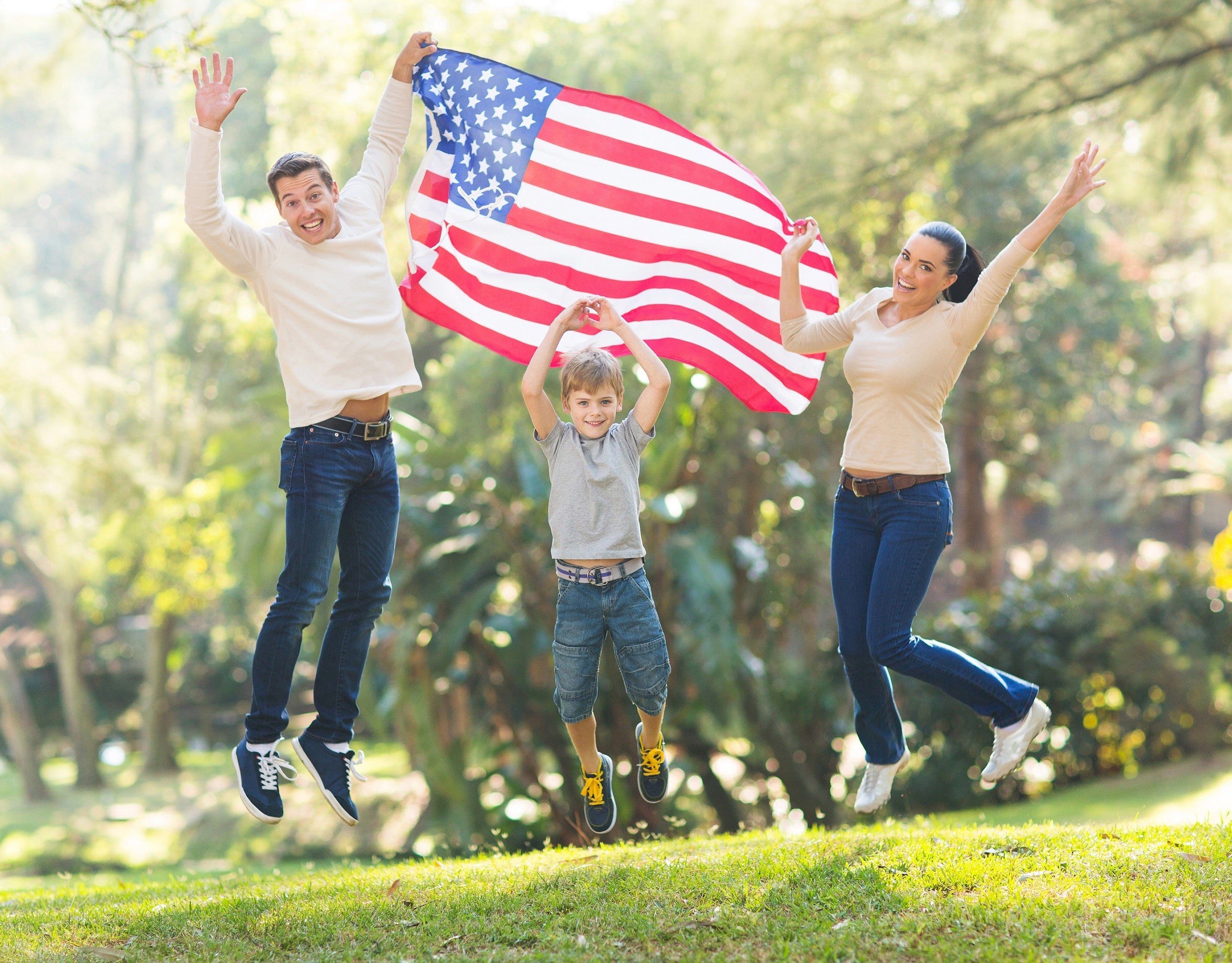 Happy family jumping outdoors while holding an American flag in a sunny park.