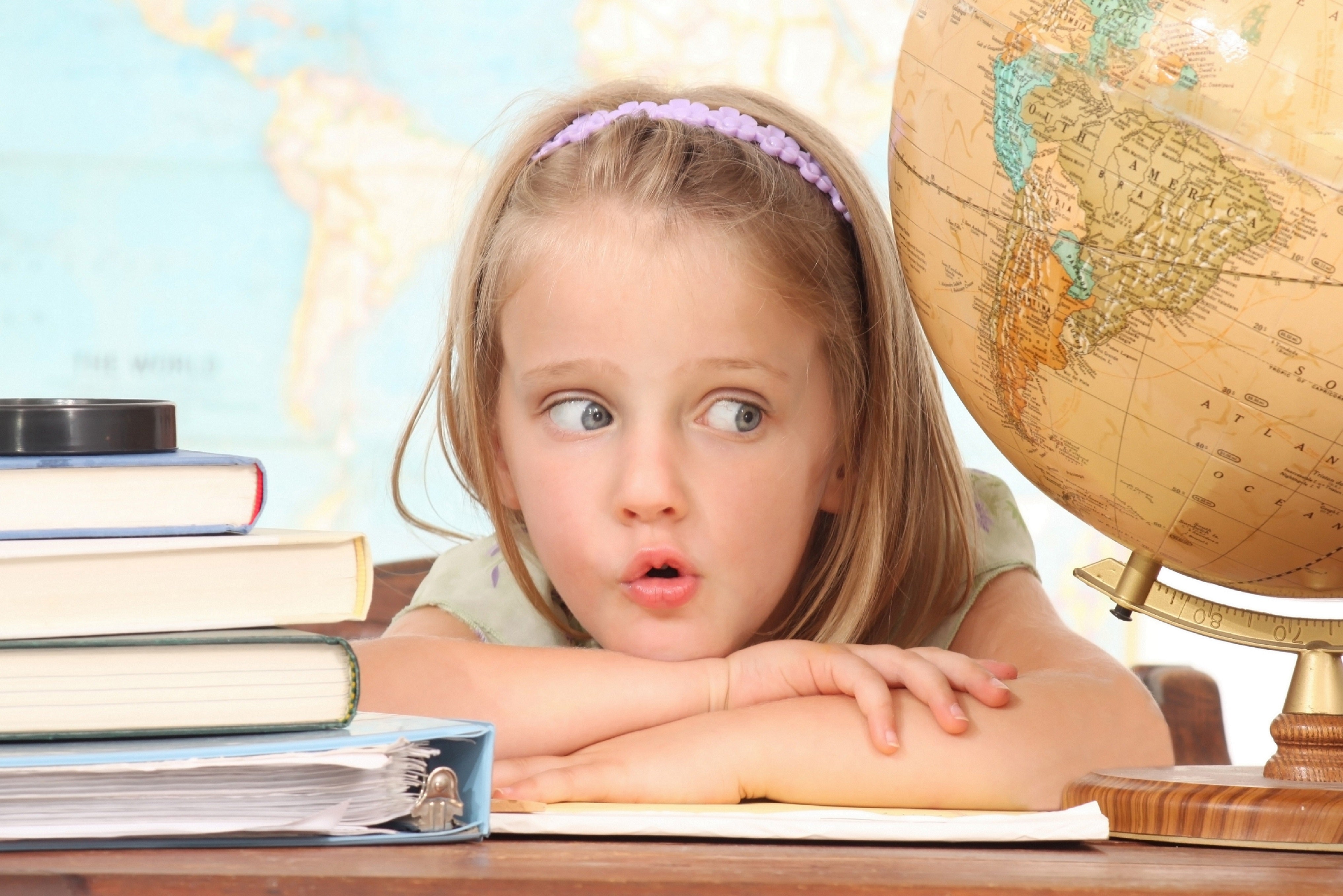 Young girl leaning on a desk beside books and a globe, looking curious in a classroom setting.