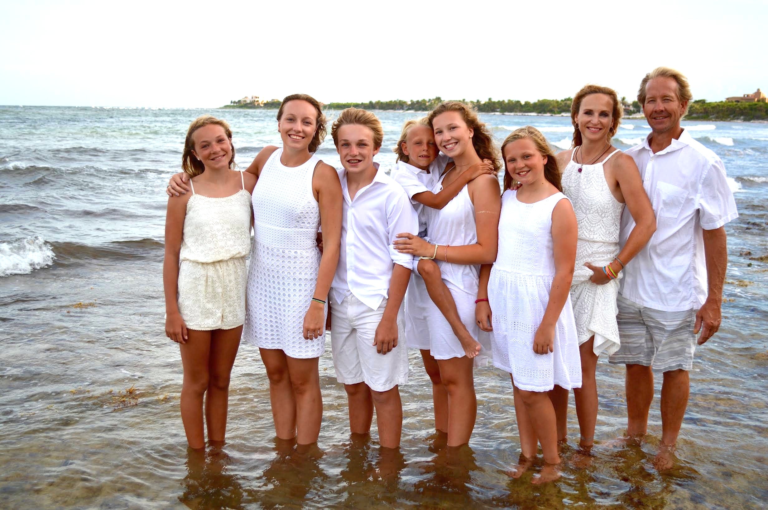 Jennifer Adams posing with her family standing together in shallow ocean water at the beach, all dressed in white outfits.
