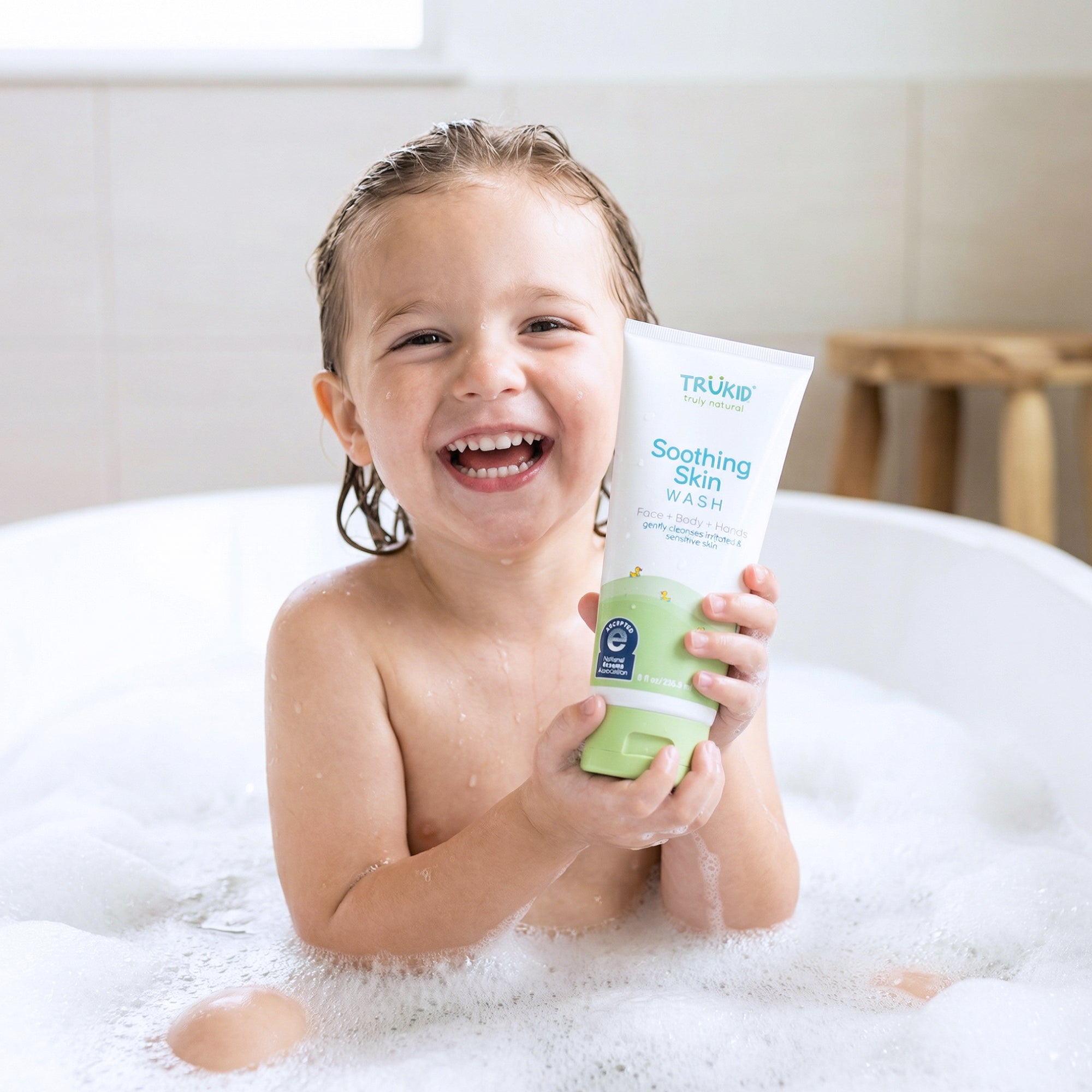 Child in a bathtub holding a tube of TruKid Soothing Skin Wash.